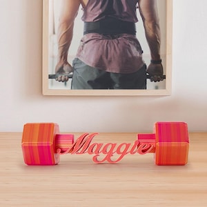 Pode incluir: Um objeto decorativo personalizado em forma de haltere com o nome "Maggie" em fonte cursiva. O haltere apresenta um gradiente de tons laranja e rosa. No fundo, uma foto emoldurada de uma pessoa levantando pesos.