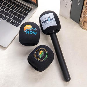 Op de afbeelding: Een zwarte microfoon met een lange zwarte handgreep en drie zwarte microfoonhoezen. Eén hoes heeft de tekst "Doing it with PURPOSE PODCAST LLC". Een andere hoes heeft de tekst "eastern sierra NOW". De derde hoes heeft de tekst "PRESENTING HOMES AUSTRALIA".