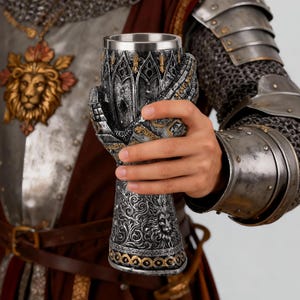 May include: A silver and gold decorative goblet with intricate medieval-style detailing. The goblet is held by a hand sculpted to resemble a gauntlet. The goblet features a stainless steel interior and ornate patterns.