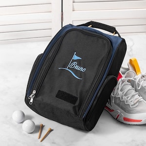 Puede incluir: Bolsa de zapatos de golf negra y azul marino con el nombre "Bruno" bordado en azul claro. La bolsa tiene un asa, cremallera y un parche de velcro. Dos pelotas de golf, dos tees y un par de zapatos de golf blancos y rojos están junto a la bolsa.