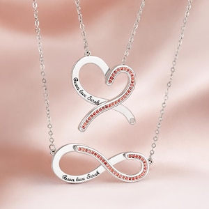 Puede incluir: Dos collares de tono plateado con símbolos de corazón e infinito. El colgante de corazón y el de infinito están grabados con el texto 'Jason love Sarah' y decorados con pequeñas piedras rojas a lo largo de un lado de cada forma.