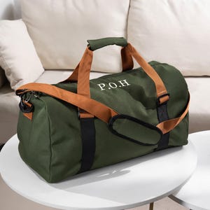 May include: An olive green duffel bag with brown straps and handles. The bag has the initials "P.O.H" in white lettering. The bag is sitting on a white table.