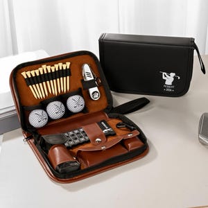Op de afbeelding: Een bruine leren golfset met ritssluiting, met golfballen, tees en een divotgereedschap. Een zwarte etui met een golferafbeelding en de tekst "ROBERT - 2026" is ook aanwezig.