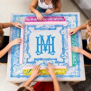 May include: A square Mahjong game table with a blue and white floral design and a large blue monogram in the center. The table is surrounded by people playing the game, with pink, green, and yellow tile racks visible.
