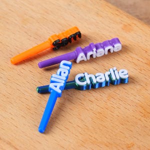 Peut inclure: Trois étiquettes nominatives personnalisées colorées avec un design unique. Les étiquettes sont orange, violettes et bleues, chacune avec un nom différent en lettres blanches. Les étiquettes sont sur une surface en bois.
