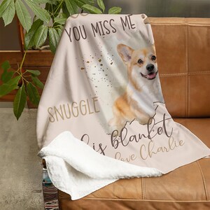 K&ouml;nnte beinhalten: Eine beige-wei&szlig;e Decke mit einem l&auml;chelnden Corgi-Hund. Auf der Decke steht der Text "You miss me", "Snuggle", "This blanket" und "Love Charlie". Die Decke ist &uuml;ber ein braunes Ledersofa drapiert.