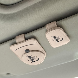 May include: Two beige car visor organizers with a personalized initial and name design. The organizers are made of leather-like material and feature a loop for hanging. The text "Louise" is visible.
