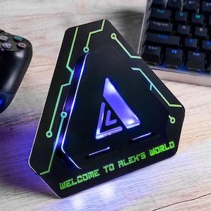 May include: Black triangular gaming accessory with green and blue neon accents. The device features a glowing blue triangle and the text "WELCOME TO ALEX'S WORLD". A game controller and keyboard are visible in the background.