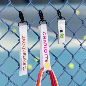 May include: Three white fabric keychains with black clips hang on a chain-link fence. One keychain says "JACQUELINE" in gold, another says "CHARLOTTE" in pink, and the third has a tennis ball graphic and the letter "B".
