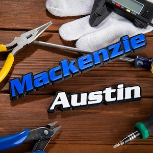 May include: Two nameplates, "Mackenzie" in blue and "Austin" in white, rest on a wooden surface with tools. The "Mackenzie" nameplate has a blue font with a black outline. The "Austin" nameplate has a white font with a black outline.