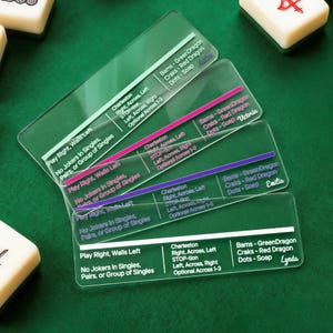 Pode incluir: Folhas de truques de Mahjong em acrílico transparente com texto impresso. Cada folha tem um sotaque de cor diferente e inclui instruções do jogo e nomes de jogadores. As folhas estão sobre uma superfície verde, com peças de Mahjong no fundo.