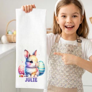Könnte beinhalten: Weißes Küchentuch mit Waffelstruktur, das eine Aquarellillustration eines Hasen, Ostereiern und dem Namen "JULIE" in Lila zeigt. Der Hase hat rosa Ohren und einen flauschigen, bunten Körper. Die Eier sind mit Mustern verziert.