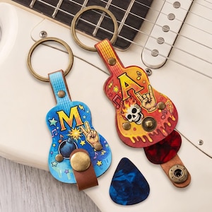 以下が含まれることがあります： ギター型のレザーキーホルダー2点。金属リングとスナップボタン付き。青い方は「M」の文字、爆弾、ピースサインが描かれています。赤と黄色のもう一方は「A」の文字、ドクロ、ハンドサインが描かれています。ギターピック2枚付き。