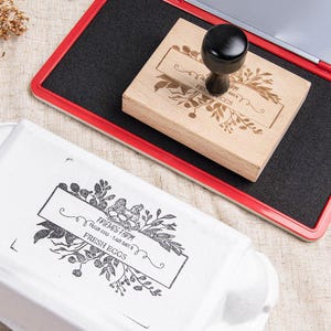 May include: A wooden stamp with a black handle and floral design, reading "FRESH EGGS". The stamp is on a red and black ink pad, with a stamped image on a white egg carton. The text on the carton reads "FRIENDS FARM FRESH EGGS".