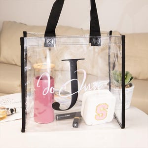 May include: A clear tote bag with black trim and handles, featuring a large black letter "J" and the words "Jo Luxe" in white script. Inside are a pink tumbler, makeup, and a white case with a pink "S".