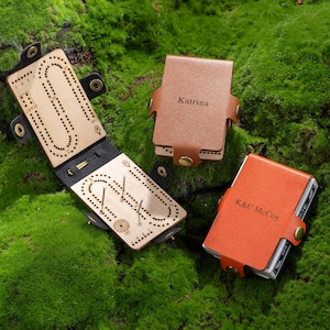 Op de afbeelding: Een houten cribbagebord met een zwarte leren hoes, open om metalen pinnen te onthullen. Er worden twee leren kaarthouders getoond, één bruin met "Katrina" en één oranje met "K&C McCoy" in reliëf.