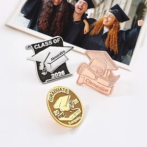 May include: Three graduation pins featuring mortarboard designs. One pin is silver with "Class of 2026" and "Alexandra" text. Another is copper-coloured with "Clementine" text. The third is gold with "Graduate 2026" and "Evangeline" text.