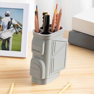 Puede incluir: Un organizador de escritorio gris con forma de bolsa de golf que contiene lápices y bolígrafos. El organizador tiene las palabras "Love you" en el lateral. También se ve una foto enmarcada y tees de golf.