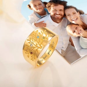 May include: A gold-colored ring with a tree design and colorful gemstones. The ring has the inscription "We love you A&F" engraved on the side. The ring is a family-themed jewelry item.