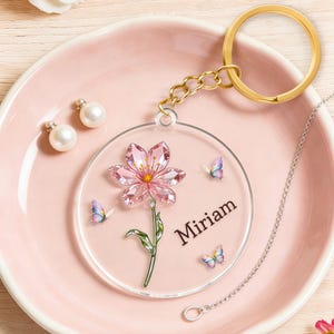 May include: A clear acrylic keychain with a pink flower design and the name "Miriam" in black. The keychain features a gold-colored ring and chain, with small butterflies and two pearl earrings on a pink plate.