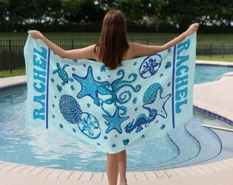 May include: A light blue beach towel featuring various sea creatures in shades of blue. The towel has the name "RACHEL" printed vertically on the side. The design includes starfish, an octopus, and a seahorse.