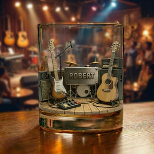 Könnte beinhalten: Ein klares Glas mit einer Miniaturbühne im Inneren, auf der zwei Gitarren, ein Schlagzeug und ein Mikrofon stehen. Die Bühne hat einen Holzboden und ein Metallschild mit der Aufschrift "ROBERT". Der Hintergrund ist verschwommen, was auf einen Konzertsaal hindeutet.