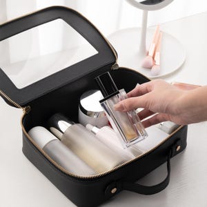 May include: Black cosmetic bag with a clear top, gold zipper, and handle. The bag is open, revealing various cosmetic bottles and jars. A hand is reaching in to grab a clear perfume bottle.
