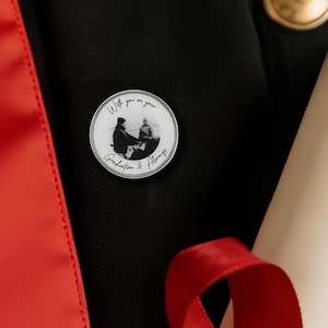 Puede incluir: Pin redondo con borde plateado, con una foto en blanco y negro de dos personas. El pin tiene las palabras "With you on your Graduation & Always" en una fuente manuscrita. Fondo negro con una cinta roja.