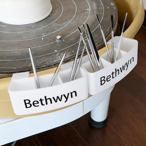 May include: White plastic tool holder with the name "Bethwyn" printed on the side, holding various pottery tools. The tools are silver and black, and the holder is positioned near a pottery wheel.