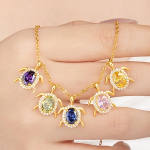 May include: A gold-colored necklace featuring five turtle pendants, each with a different colored oval gemstone. The gemstones are surrounded by small, clear stones. The turtle pendants are connected by a delicate chain.