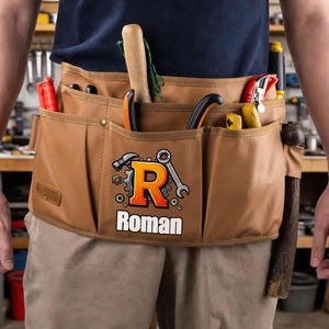 May include: A brown canvas tool belt with multiple pockets, holding various tools like a hammer, pliers, and screwdrivers. The belt features the name "Roman" with a logo of a hammer, wrench, and gear.