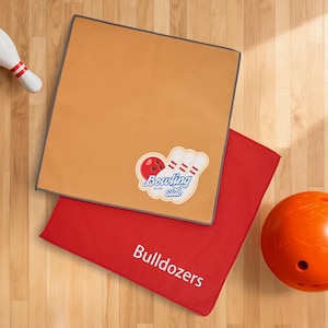 Op de afbeelding: Twee bowlinghanddoeken, één bruin en één rood, worden getoond op een houten oppervlak. De bruine handdoek heeft een "Bowling Club" logo. Op de rode handdoek staat het woord "Bulldozers". Een oranje bowlingbal en een witte bowlingpin zijn ook zichtbaar.