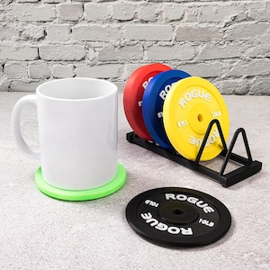 May include: A white ceramic mug rests on a green coaster. A set of red, blue, and yellow weight plates are in a black metal rack. A black weight plate coaster with "ROGUE" text is also present. The weight plates are labeled "10LB" and "15LB".