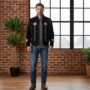 May include: A black suede jacket featuring long fringe along the sleeves and chest, accented with white leather lacing and circular beaded medallions on the chest and sleeves. The jacket is worn over a grey t-shirt and blue denim jeans.