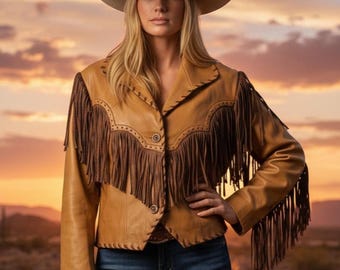 Op de afbeelding: Een bruinleren jasje met bruine franjes en een bijpassende cowboyhoed. De jas heeft decoratieve stiksels en knoopsluitingen. De persoon draagt een blauwe spijkerbroek. De achtergrond toont een zonsondergang boven een woestijnlandschap.