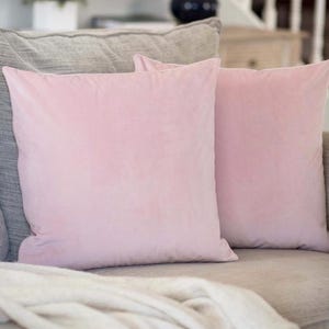 May include: Two square, light pink velvet throw pillows rest on a gray sofa, with a folded white blanket in the foreground. The pillows have a soft, plush appearance, and the overall scene suggests comfort and relaxation. A decorative vase is visible in the background.