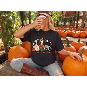 Puede incluir: Una camiseta negra con un gráfico de tres patos disfrazados de Halloween: uno de momia, uno de espantapájaros y uno de esqueleto. El diseño incluye pequeños murciélagos, fantasmas y una calabaza de Halloween impresos en la parte delantera.