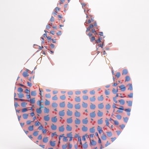 May include: A crescent-shaped shoulder bag in light pink featuring a repeating pattern of blue apples and red cherries. The bag has a ruffled trim along the bottom edge, a gathered scrunchie-style strap with small bows, and a gold zipper with a beaded pull.