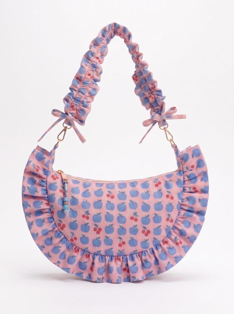 May include: A crescent-shaped shoulder bag in light pink featuring a repeating pattern of blue apples and red cherries. The bag has a ruffled trim along the bottom edge, a gathered scrunchie-style strap with small bows, and a gold zipper with a beaded pull.