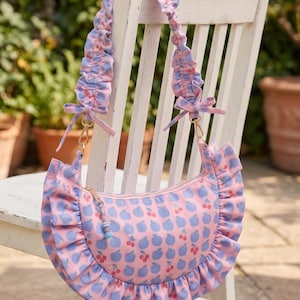 May include: A crescent-shaped shoulder bag featuring a pink background with a repeating pattern of blue apples and red cherries. The bag has a ruffled trim along the bottom edge and a matching ruffled shoulder strap with small bows at the ends.