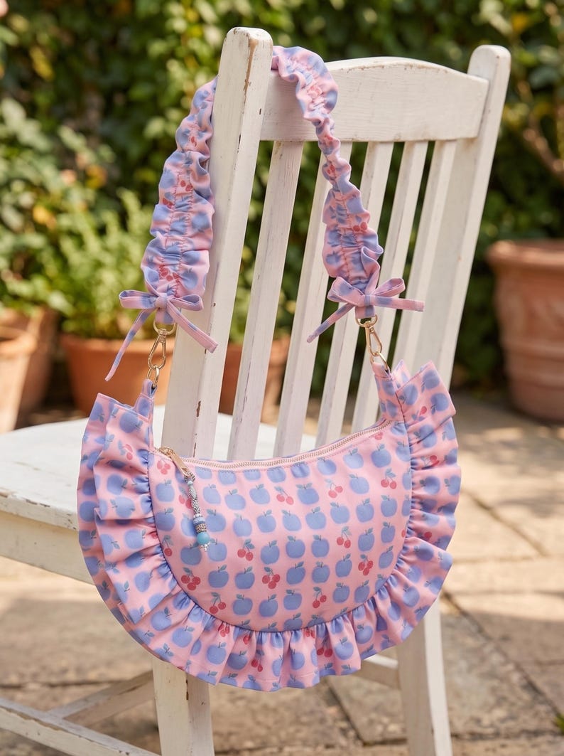May include: A crescent-shaped shoulder bag featuring a pink background with a repeating pattern of blue apples and red cherries. The bag has a ruffled trim along the bottom edge and a matching ruffled shoulder strap with small bows at the ends.