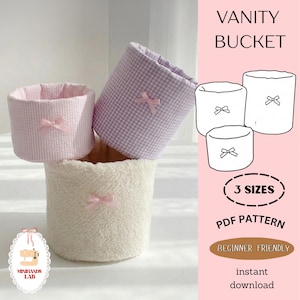 Pode incluir: Três baldes de vaidade em texturas rosa, xadrez lavanda e sherpa creme, cada um adornado com um laço rosa. A imagem inclui um desenho de linha dos baldes e o texto "VANITY BUCKET", "3 SIZES", "PDF PATTERN" e "BEGINNER FRIENDLY".