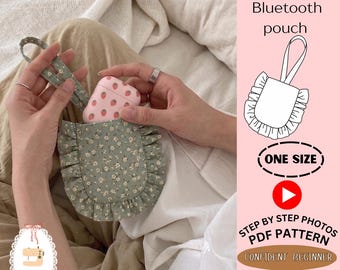 Peut inclure: Une pochette en tissu vert sauge avec un petit motif floral blanc et des bords à volants, dotée d'une dragonne assortie. La pochette contient un étui rose à motifs fraises. Le texte indique Ruffled Bluetooth pouch, ONE SIZE, STEP BY STEP PHOTOS PDF PATTERN, CONFIDENT BEGINNER et MINHANDS LAB.