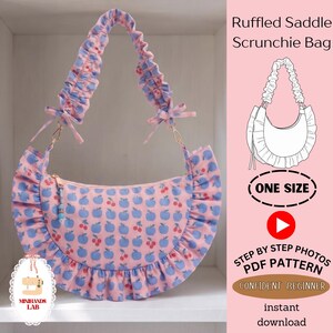 May include: A pink saddle bag with a repeating blue apple and red cherry pattern, featuring a ruffled edge and a gathered scrunchie-style shoulder strap with small side bows. The bag includes a zipper closure with a small beaded pull tab.