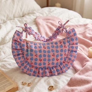 May include: A crescent-shaped handbag in light pink featuring a repeating pattern of blue apples and red cherries. The bag has a ruffled trim along the outer edge, a gathered scrunchie-style handle with tied ends, and a small beaded zipper pull.