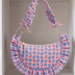 May include: A crescent-shaped shoulder bag in light pink fabric featuring a repeating pattern of blue apples and small red cherries. The bag has a ruffled trim along the bottom edge, a gathered scrunchie-style strap, and a gold zipper with a bead charm.