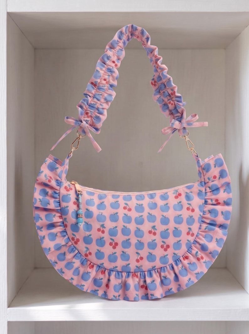 May include: A crescent-shaped shoulder bag in light pink fabric featuring a repeating pattern of blue apples and small red cherries. The bag has a ruffled trim along the bottom edge, a gathered scrunchie-style strap, and a gold zipper with a bead charm.