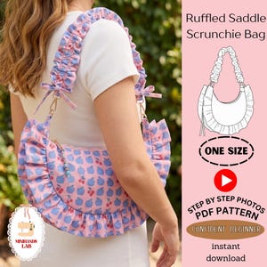 May include: A pink shoulder bag with a blue apple and red cherry print, featuring a ruffled trim and a gathered scrunchie-style strap. The bag includes a zipper closure with a beaded pull. Text on the image reads Ruffled Saddle Scrunchie Bag.