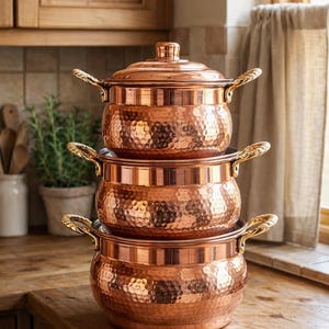 Op de afbeelding: Een stapel van drie koperen kookpotten met deksels en sierlijke handvatten. De potten hebben een gehamerde textuur en staan op een houten aanrecht. De keuken is te zien op de achtergrond.