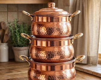 Può includere: Una pila di tre pentole di rame con coperchi e manici decorati. Le pentole hanno una texture martellata e sono esposte su un piano di lavoro in legno. L'ambiente è una cucina.